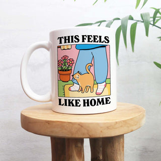 Mug with a cat illustration and text 'This feels like home' on a wooden stool.
